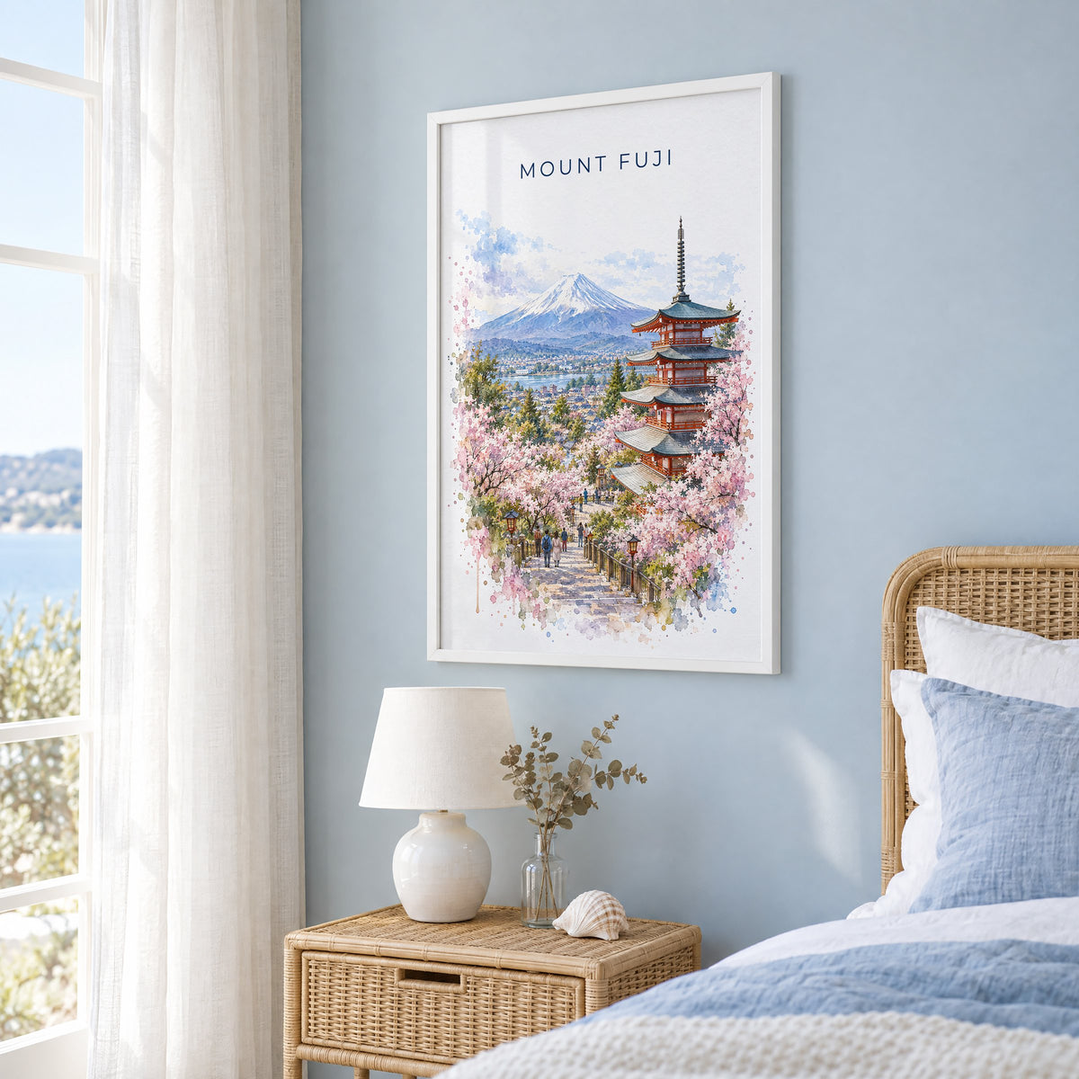 Mount Fuji watercolor print in a white frame on a pale blue wall beside a rattan bed in a coastal bedroom
