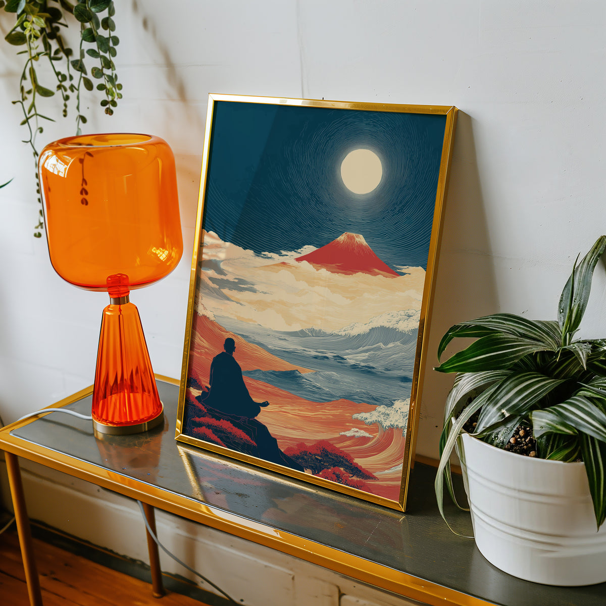 Meditating monk and red Fuji print shown in a gold frame on a console beside an orange glass lamp