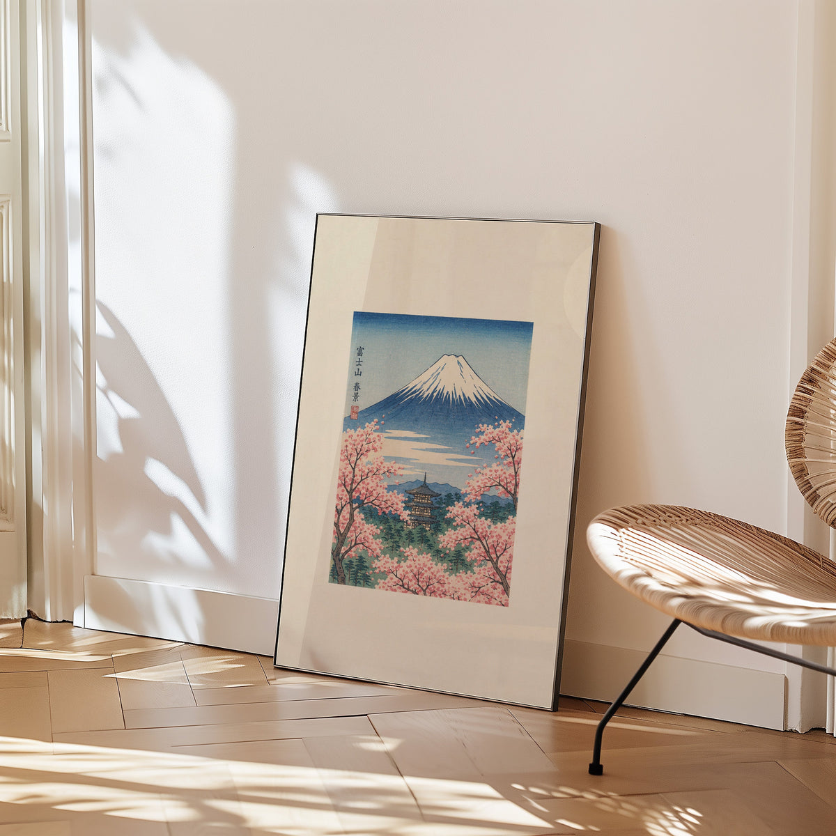 Mount Fuji with pagoda in spring print in a black frame leaning on a herringbone floor beside a woven rattan chair