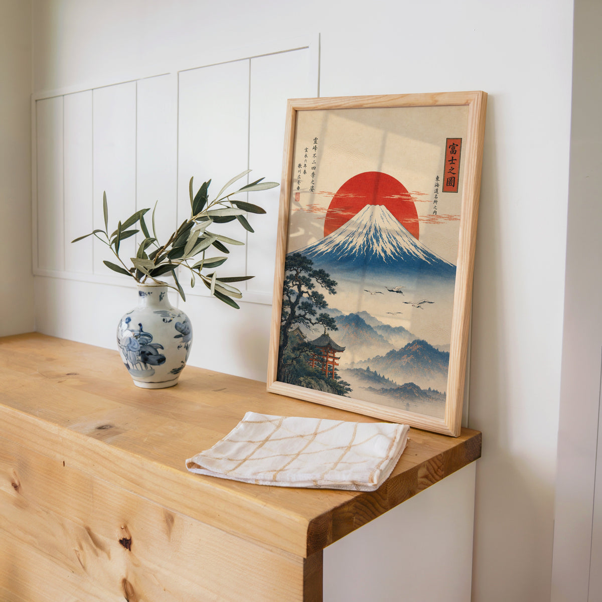 Mount Fuji with red sun and flying cranes print in a pine frame on a wooden counter beside a blue and white porcelain vase