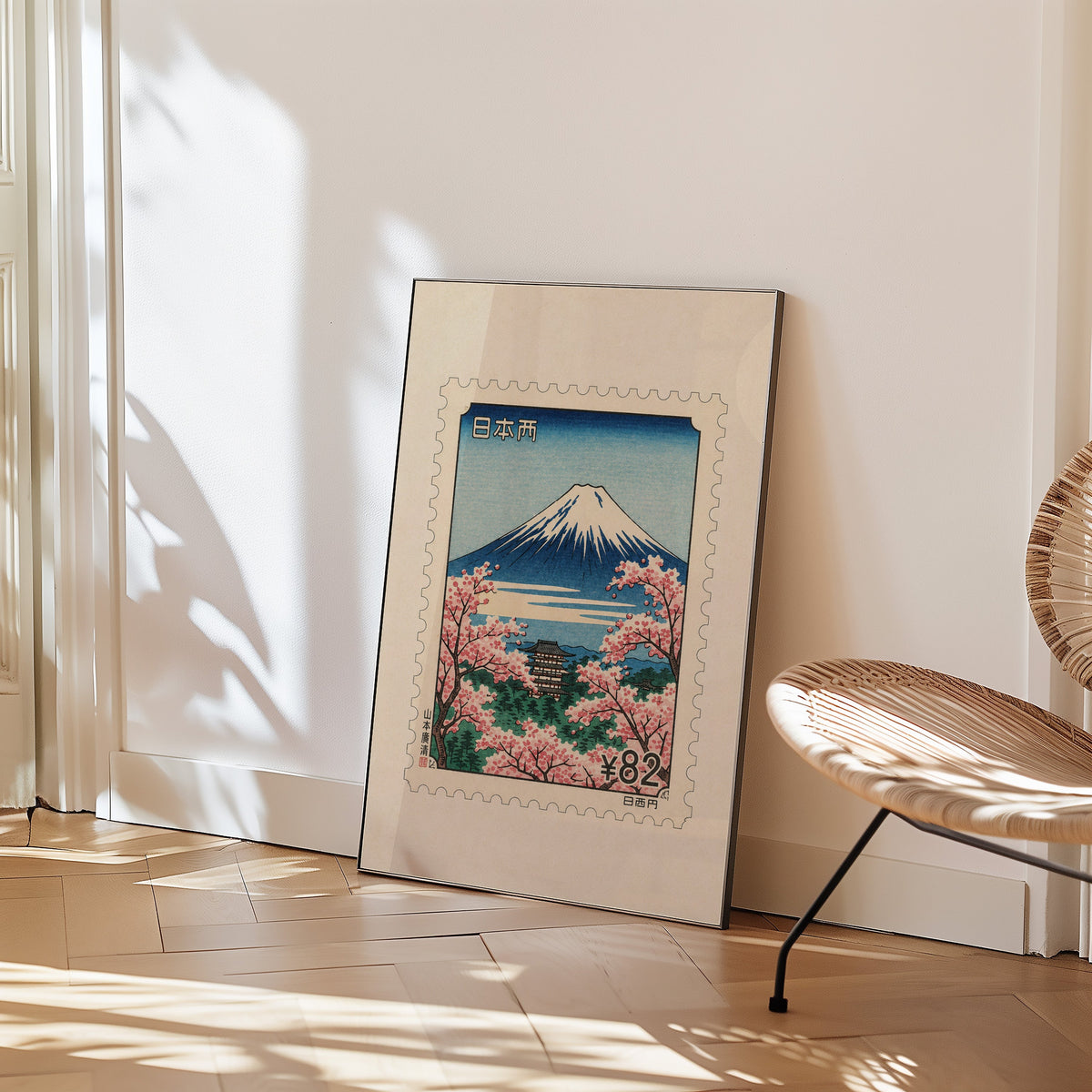 Mount Fuji sakura postage stamp print in a black frame leaning on a herringbone floor beside a woven rattan chair