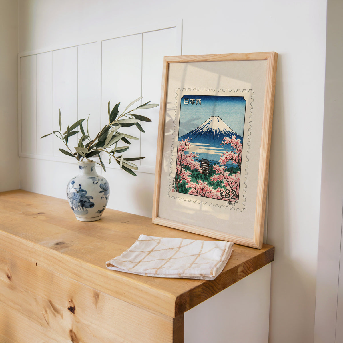 Mount Fuji sakura postage stamp print in a pine frame on a wooden counter beside a blue and white porcelain vase