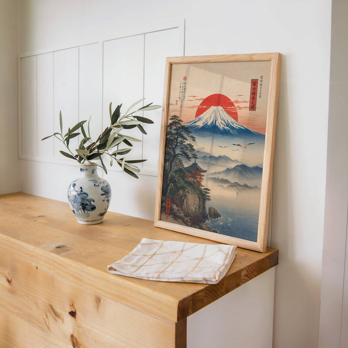 Mount Fuji sunrise with cliff shrine print in a pine frame on a wooden counter beside a blue and white porcelain vase