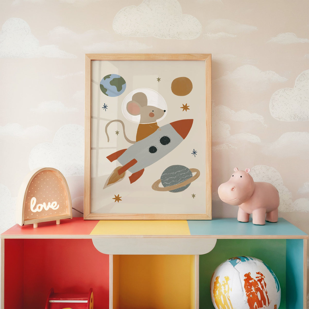 Mouse astronaut riding rocket nursery print in an oak frame on a rainbow toy shelf against cloud wallpaper