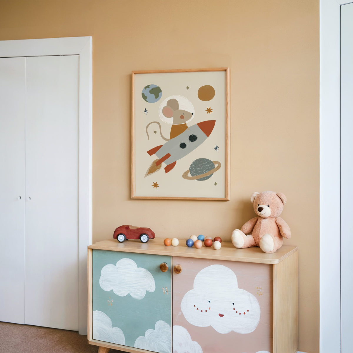 Mouse astronaut riding rocket nursery print in an oak frame on a peach wall above a cloud-painted cabinet