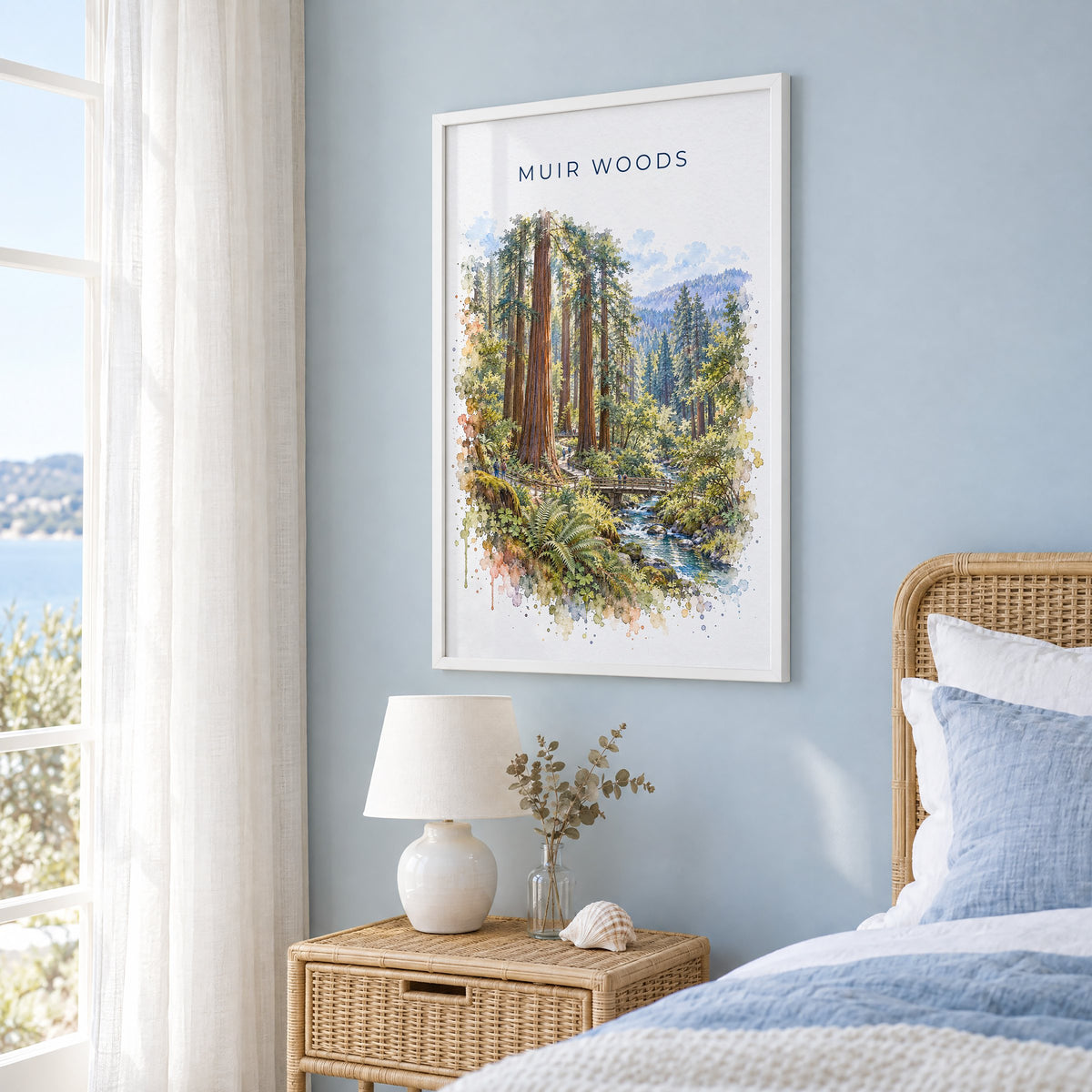 Muir Woods watercolor print in a white frame on a pale blue wall beside a rattan bed in a coastal bedroom