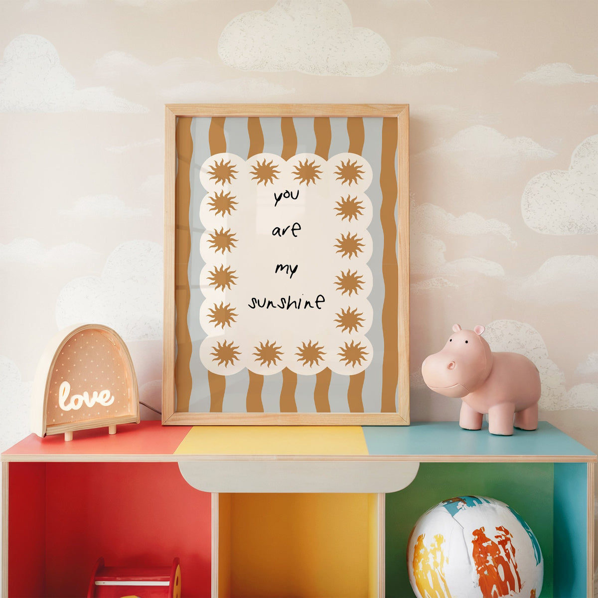 My sunshine sun border nursery print in an oak frame on a rainbow toy shelf against cloud wallpaper