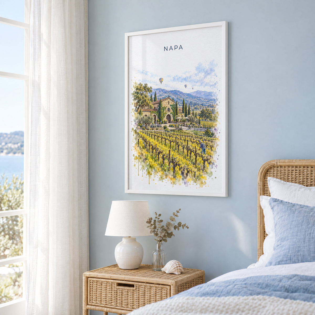Napa watercolor print in a white frame on a pale blue wall beside a rattan bed in a coastal bedroom