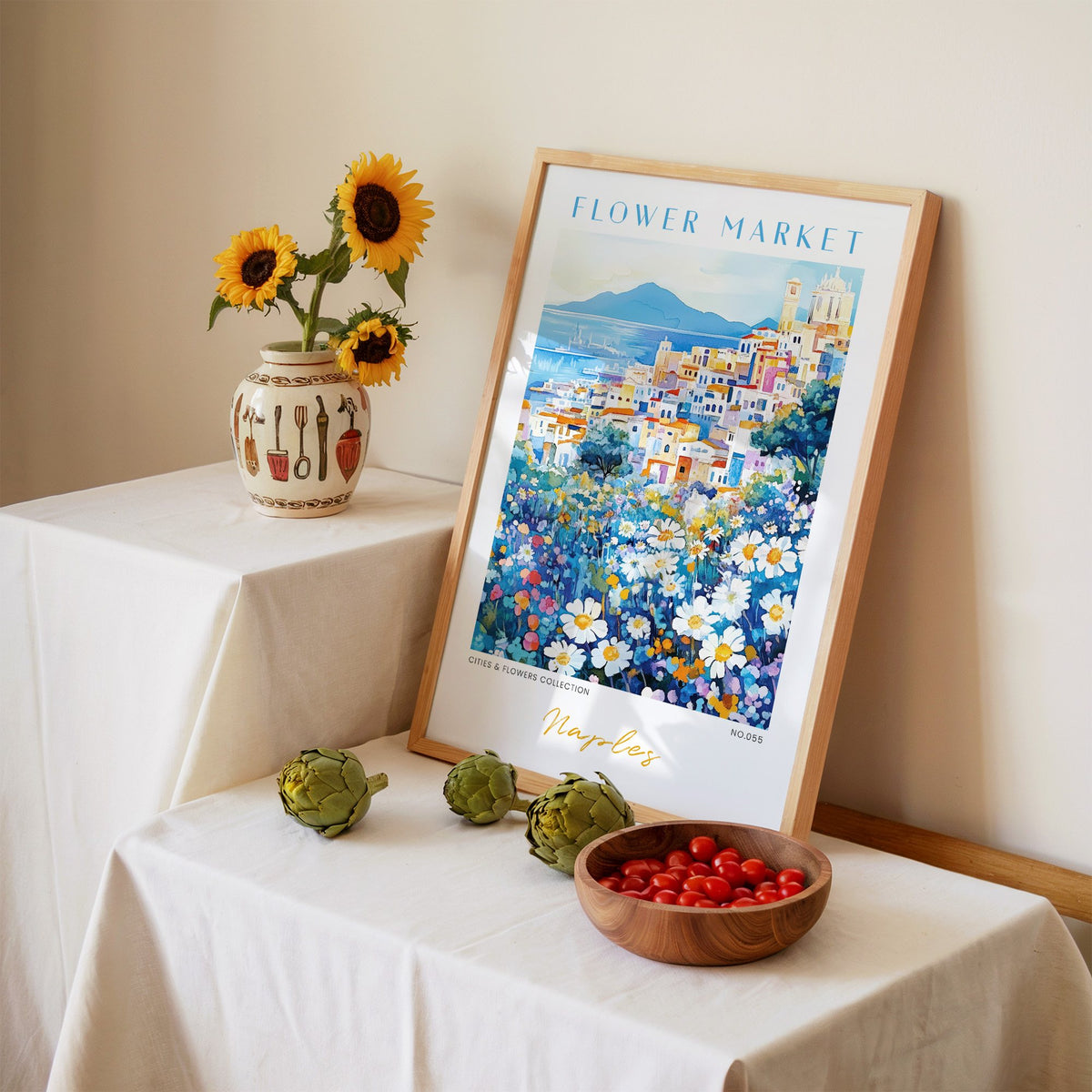 Naples flower market print in an oak frame leaning on a linen-draped table beside a vase of sunflowers