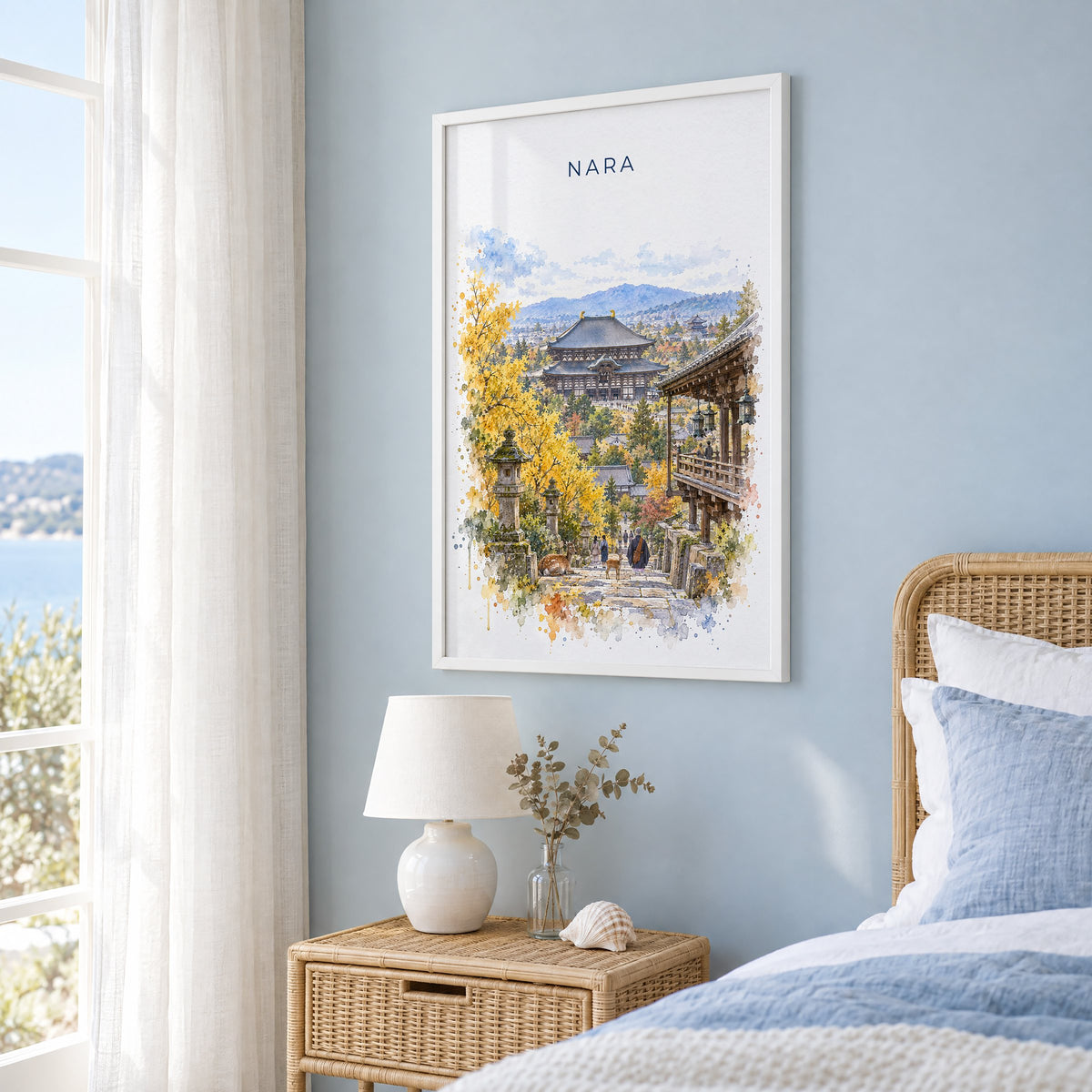 Nara watercolor print in a white frame on a pale blue wall beside a rattan bed in a coastal bedroom