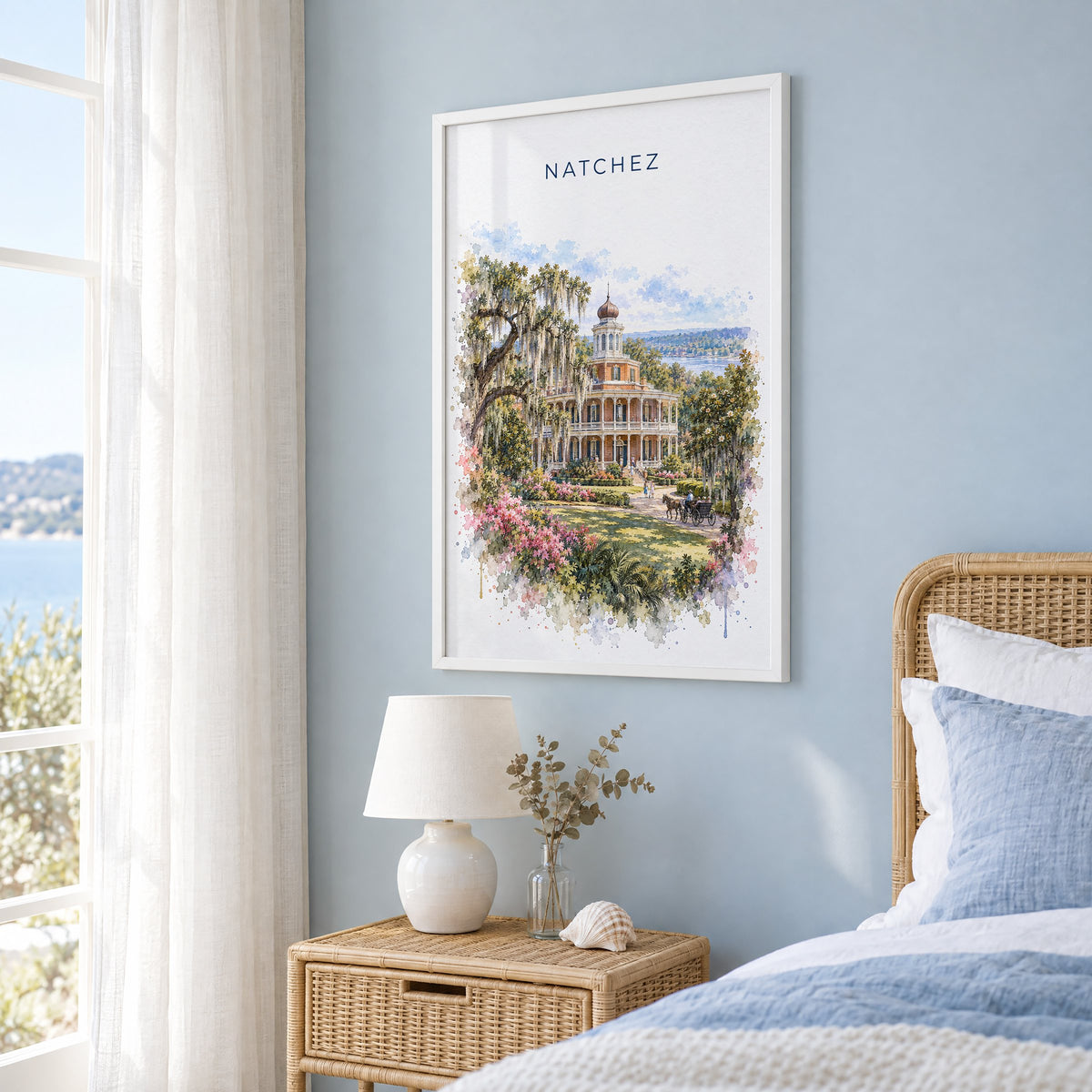 Natchez watercolor print in a white frame on a pale blue wall beside a rattan bed in a coastal bedroom