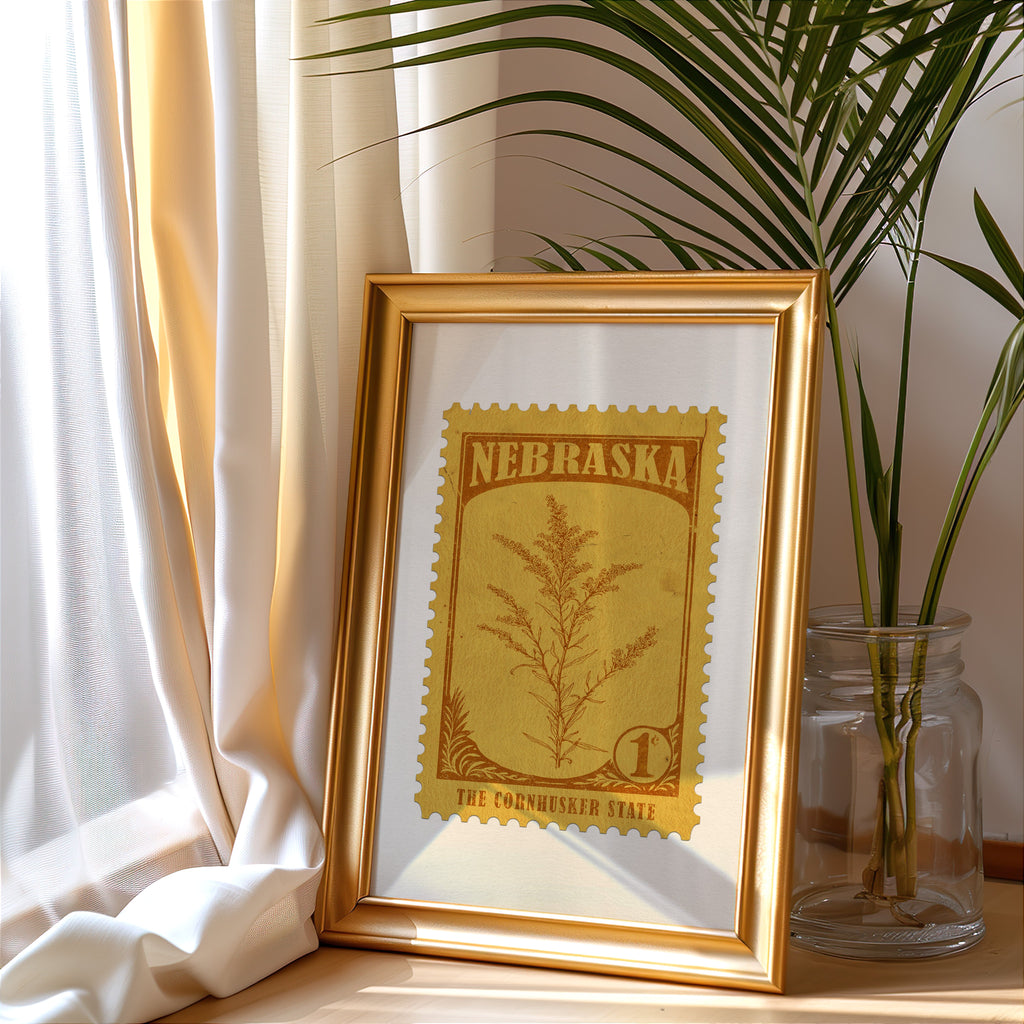 Nebraska stamp print in a gold frame leaning by a cream curtain with a palm in a glass jar