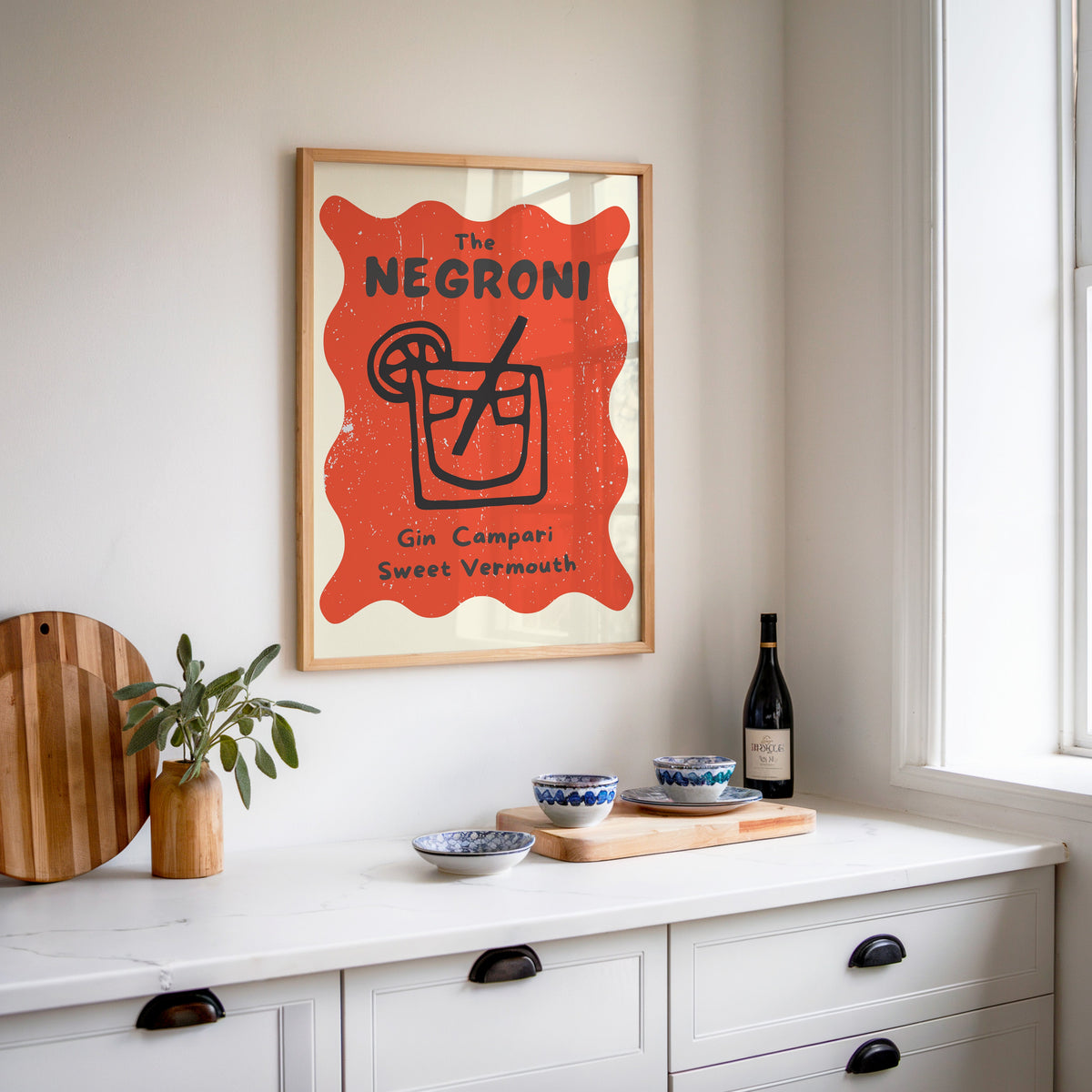 Negroni print in an oak frame above a white sideboard with a bottle of wine