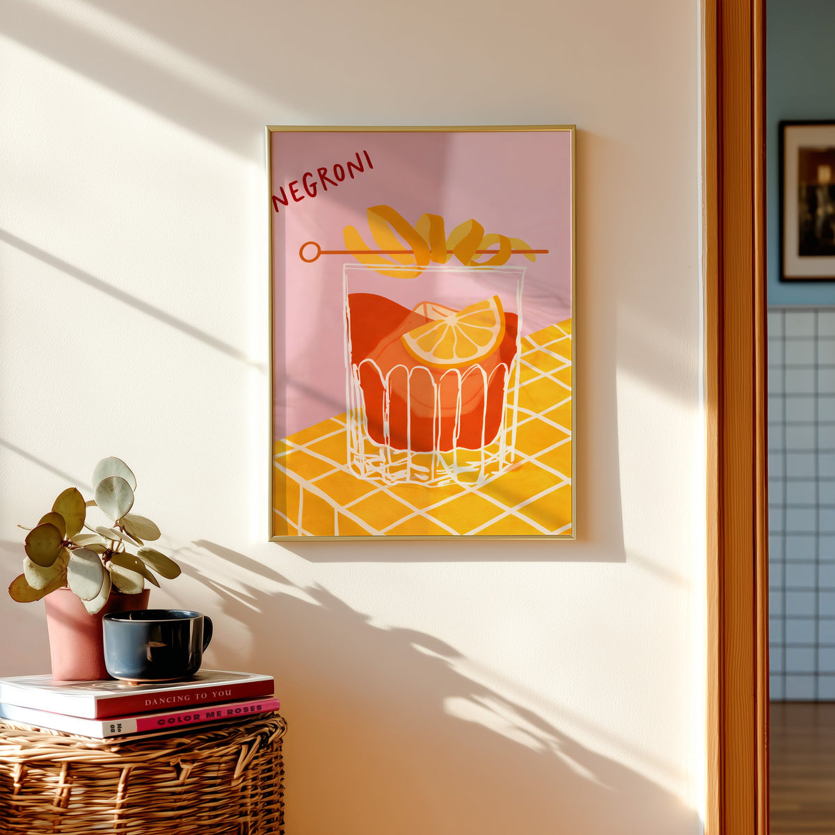 Negroni print in a gold frame above a basket of books and a pilea plant