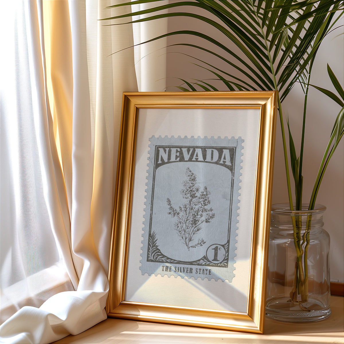 Nevada stamp print in a gold frame leaning by a cream curtain with a palm in a glass jar