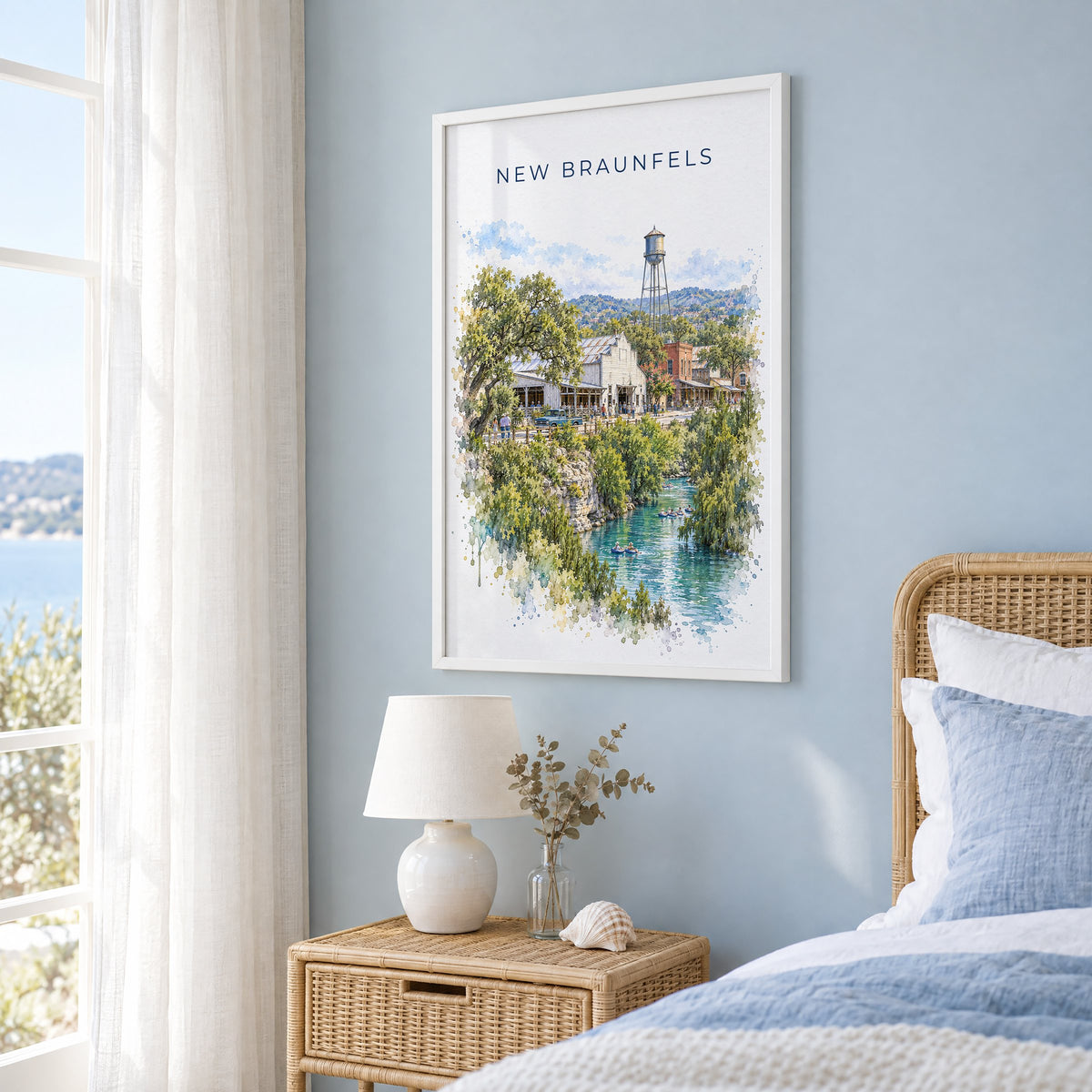 New Braunfels watercolor print in a white frame on a pale blue wall beside a rattan bed in a coastal bedroom