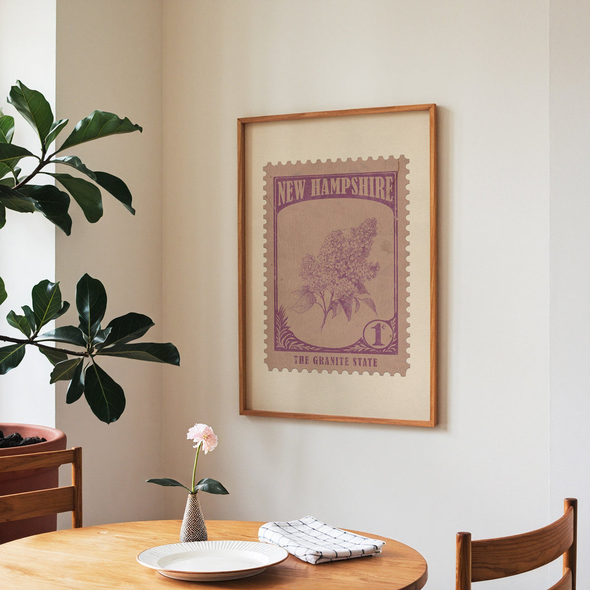 New Hampshire state flower print framed above a round wooden table with a single bloom in a vase and books