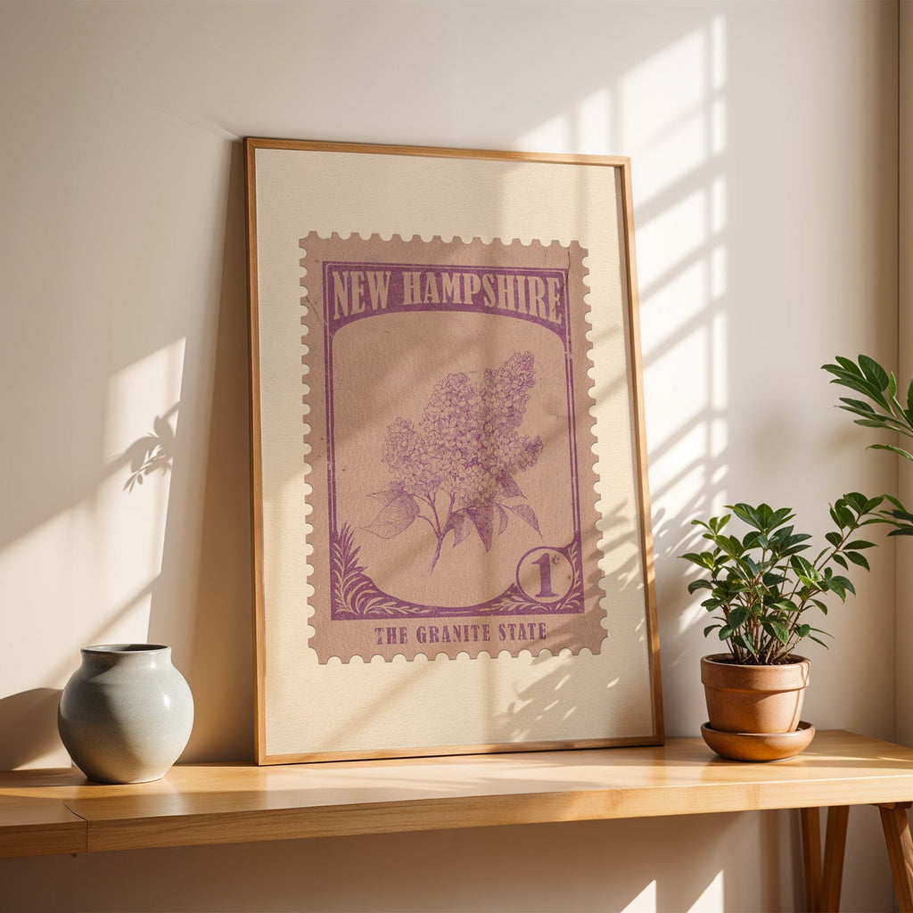 New Hampshire purple lilac stamp print in a light oak frame leaning on a wooden table beside a vase