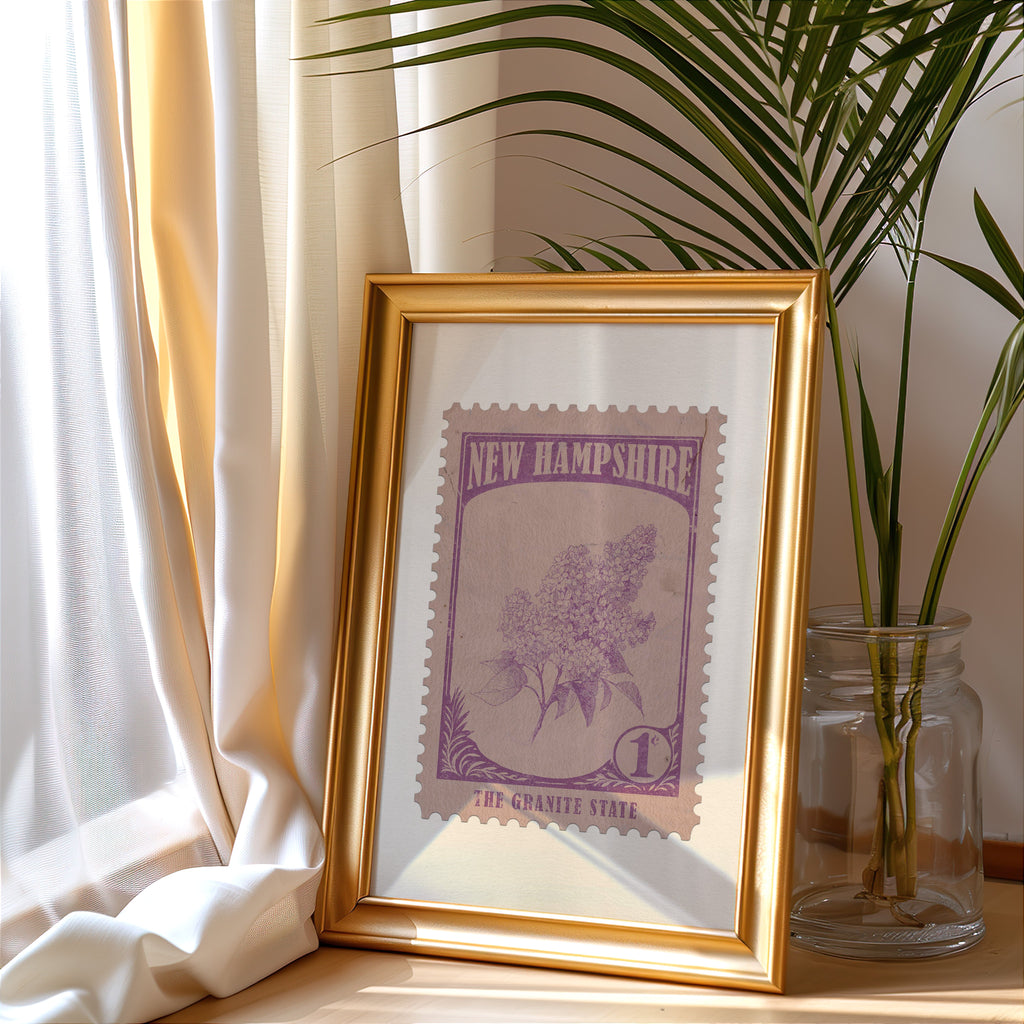 New Hampshire stamp print in a gold frame leaning by a cream curtain with a palm in a glass jar