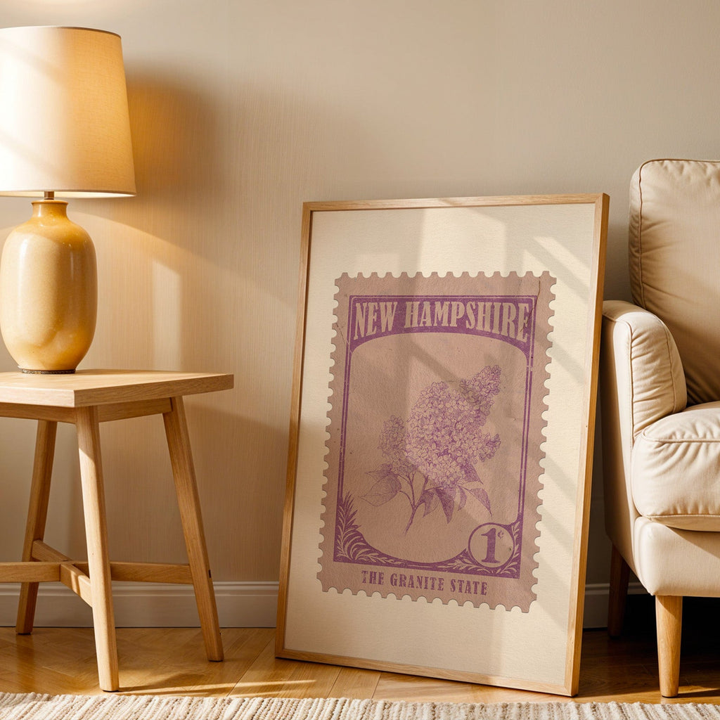 New Hampshire stamp print in a wood frame resting on the floor beside a beige armchair and a yellow lamp