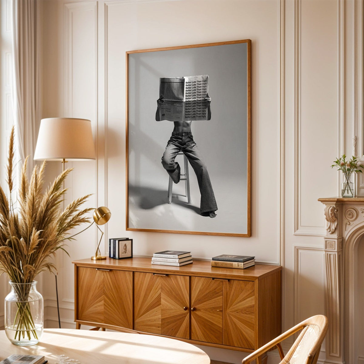 Figure on stool behind newspaper with repeated quote print in an oak frame above a sunburst wood sideboard with pampas grass