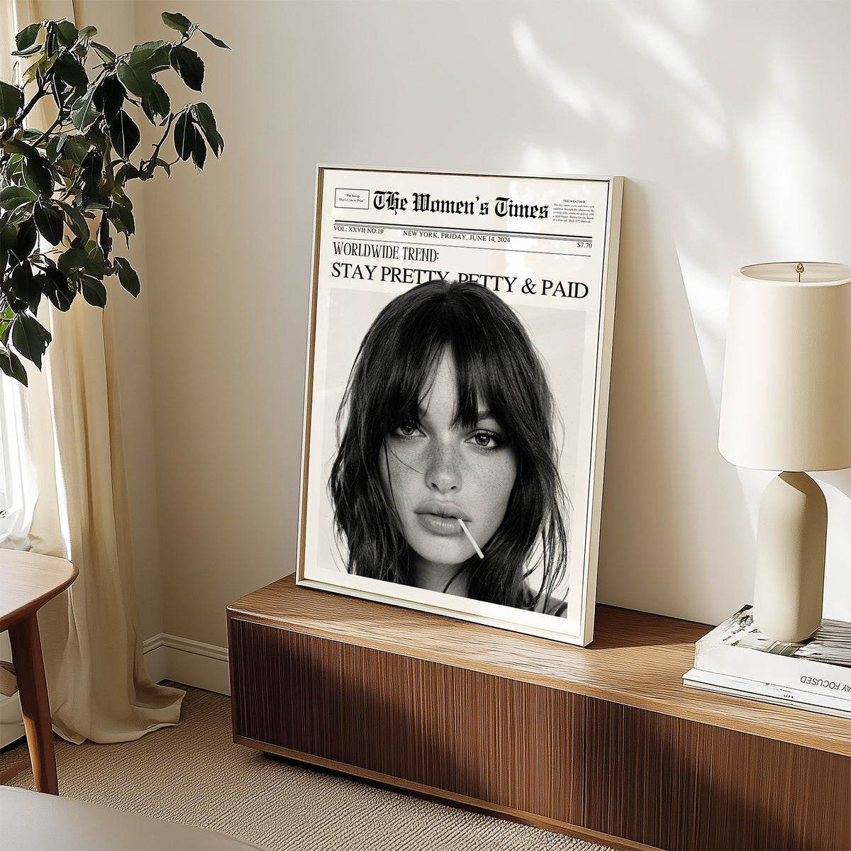 Mock front page with close-up of woman and lollipop print in a white frame on a walnut sideboard beside a ceramic lamp