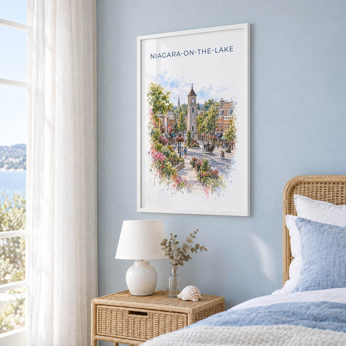 Niagara-on-the-Lake watercolor print in a white frame on a pale blue wall beside a rattan bed in a coastal bedroom