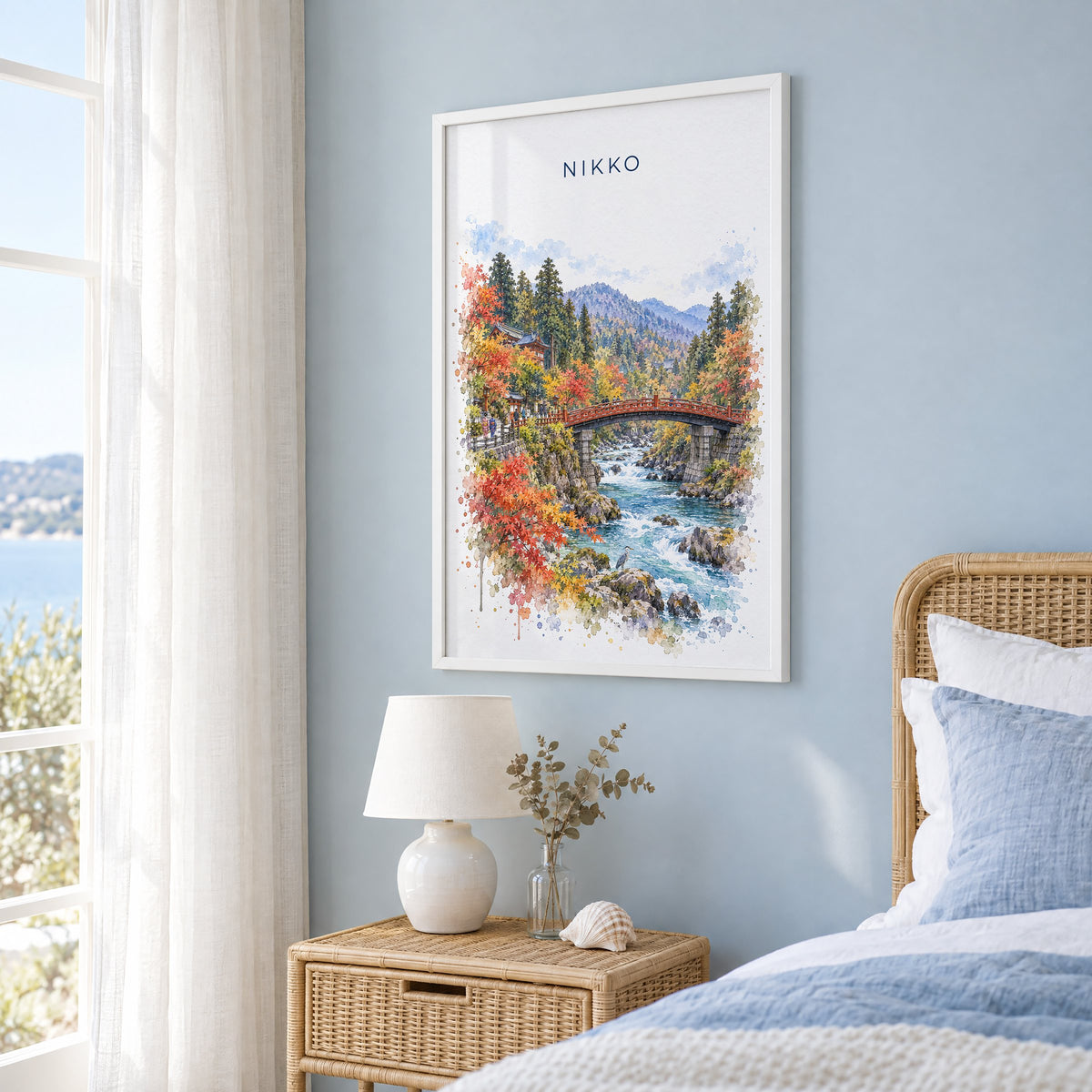 Nikko watercolor print in a white frame on a pale blue wall beside a rattan bed in a coastal bedroom