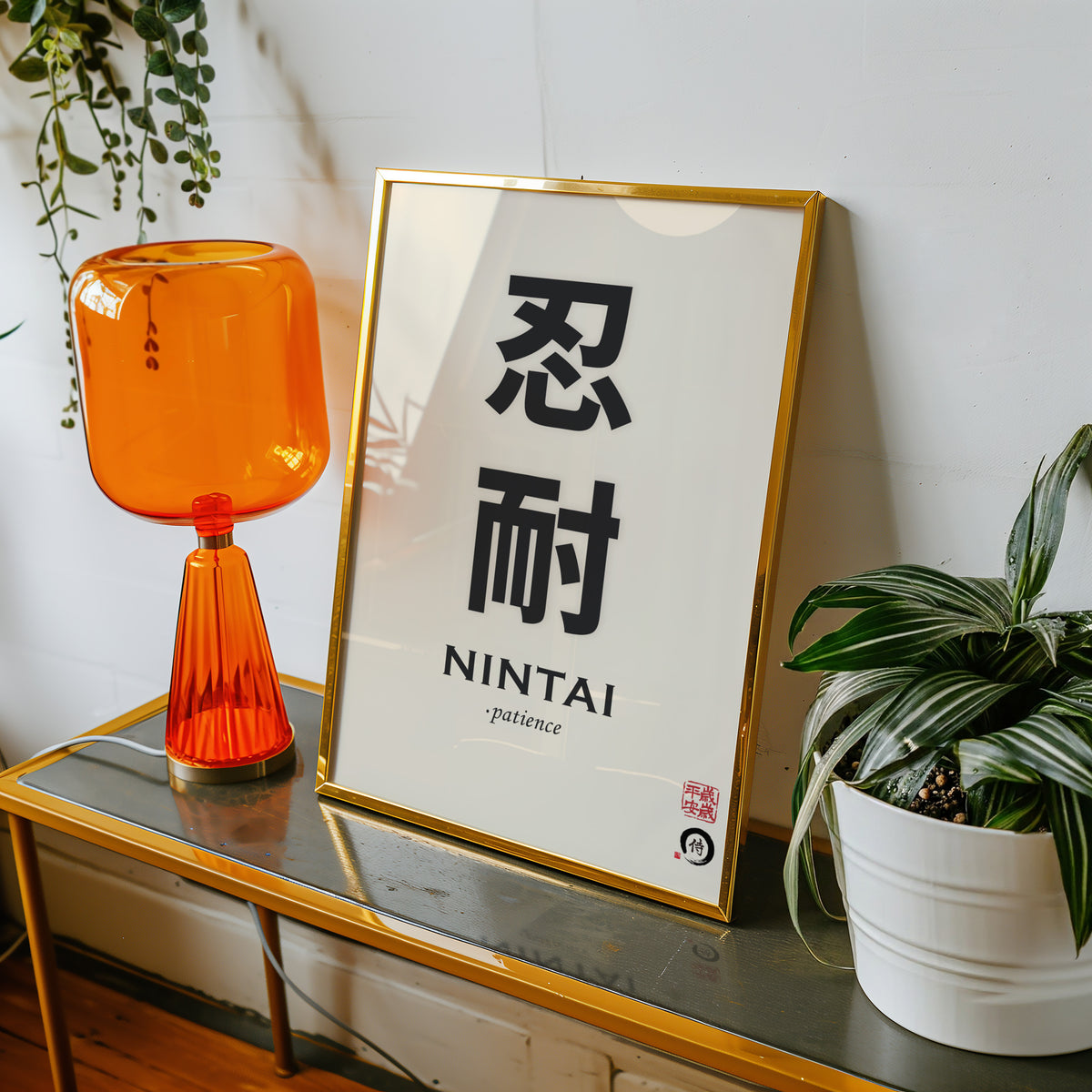 Nintai patience kanji print shown in a gold frame on a console beside an orange glass lamp
