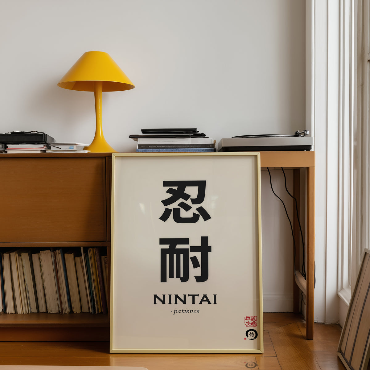 Nintai patience kanji print shown in a gold frame below a teak desk with a yellow lamp
