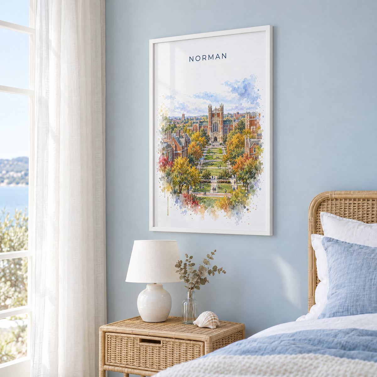 Norman, Oklahoma watercolor print in a white frame on a pale blue wall beside a rattan bed in a coastal bedroom