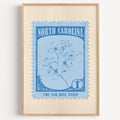 North Carolina state flower stamp print in mid blue on sky, light oak frame on a plain white wall