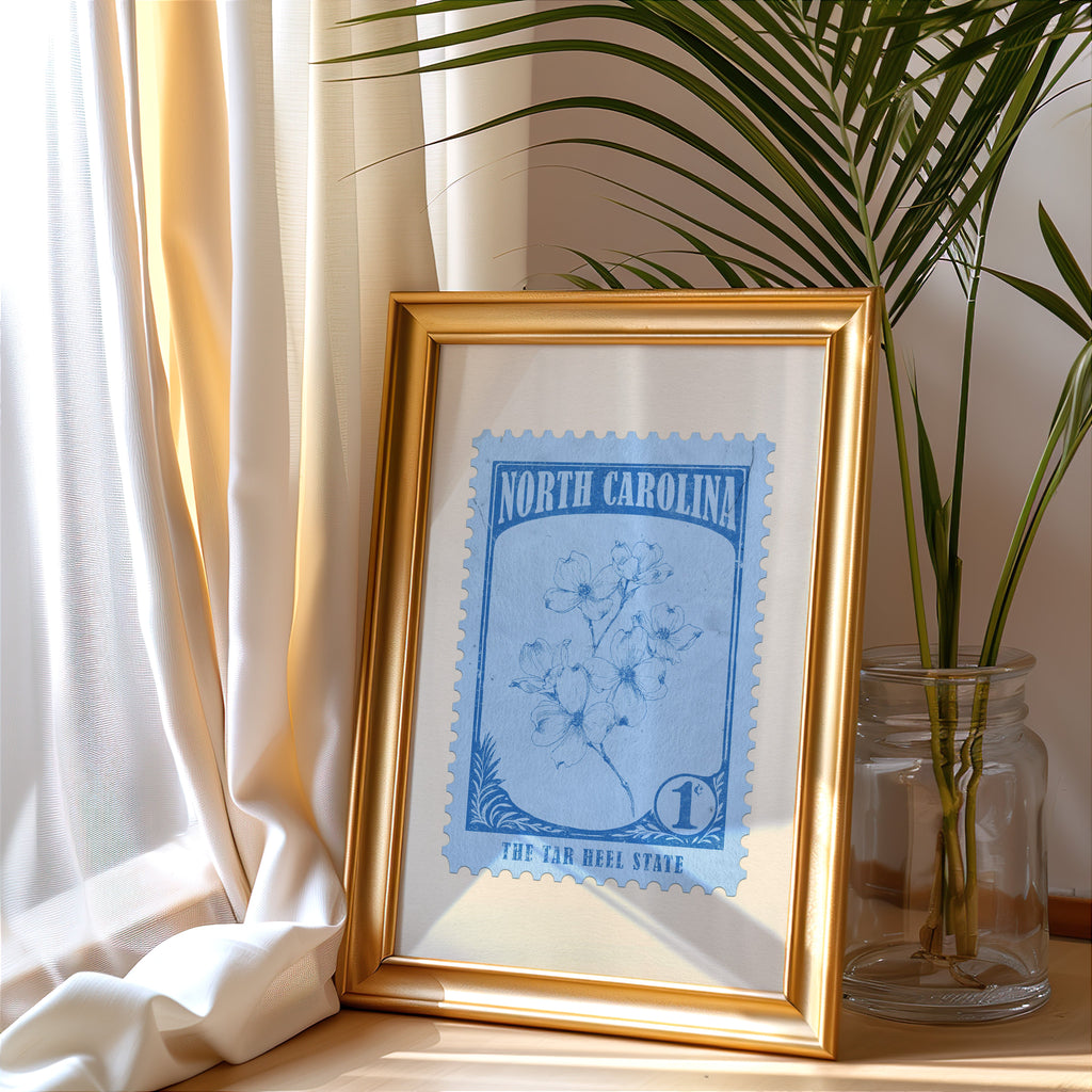 North Carolina stamp print in a gold frame leaning by a cream curtain with a palm in a glass jar