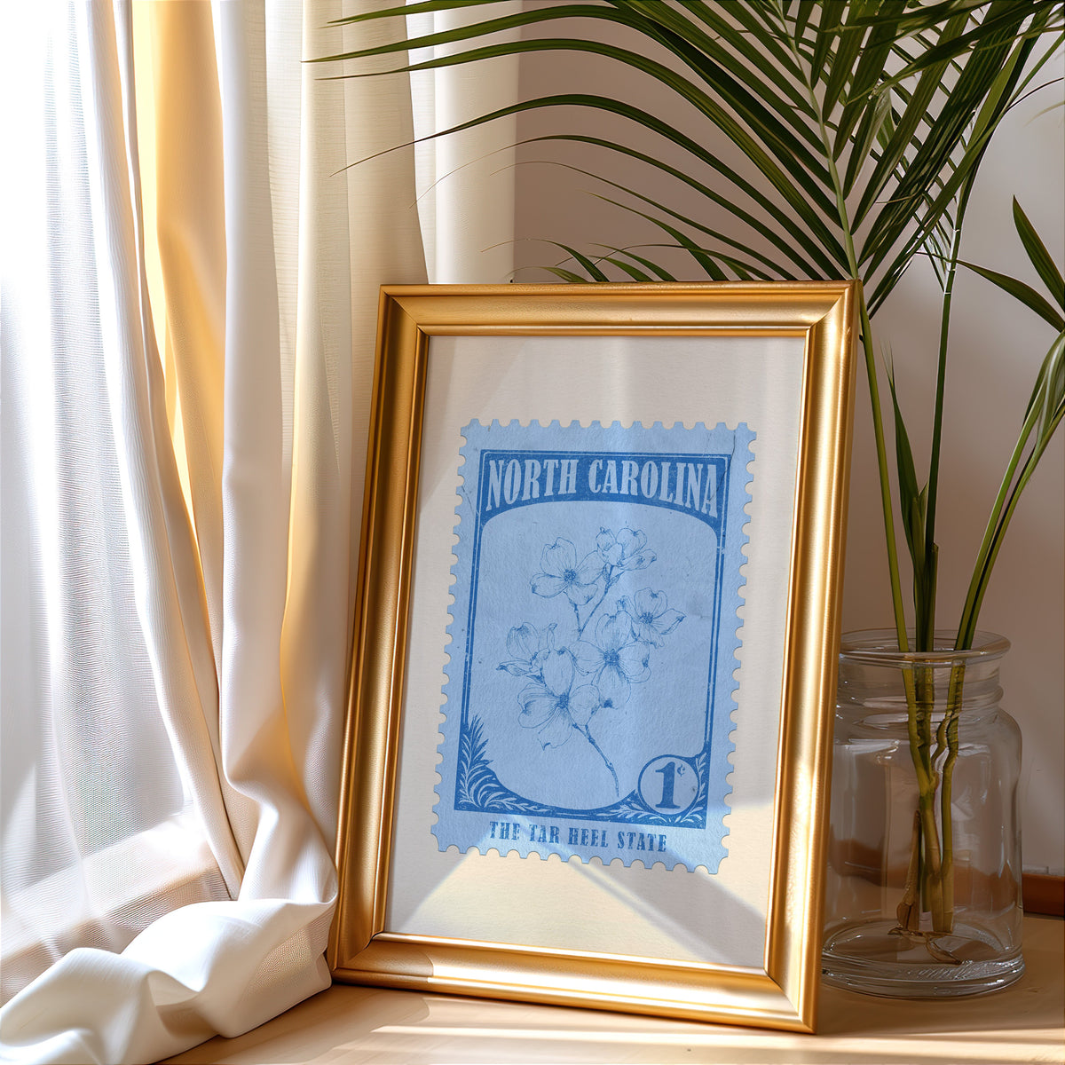North Carolina stamp print in a gold frame leaning by a cream curtain with a palm in a glass jar