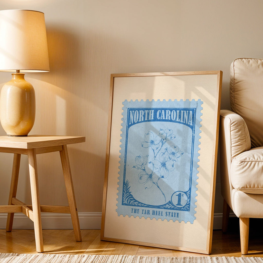 North Carolina stamp print in a wood frame resting on the floor beside a beige armchair and a yellow lamp