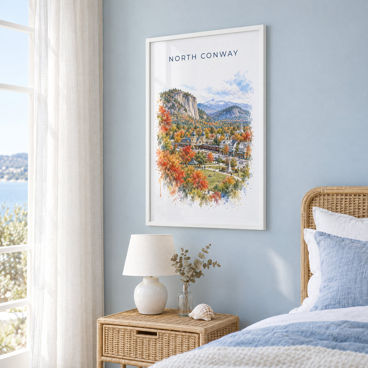 North Conway, New Hampshire watercolor print in a white frame on a pale blue wall beside a rattan bed in a coastal bedroom