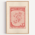 North Dakota state flower stamp print in brick red on blush, light oak frame on a plain white wall