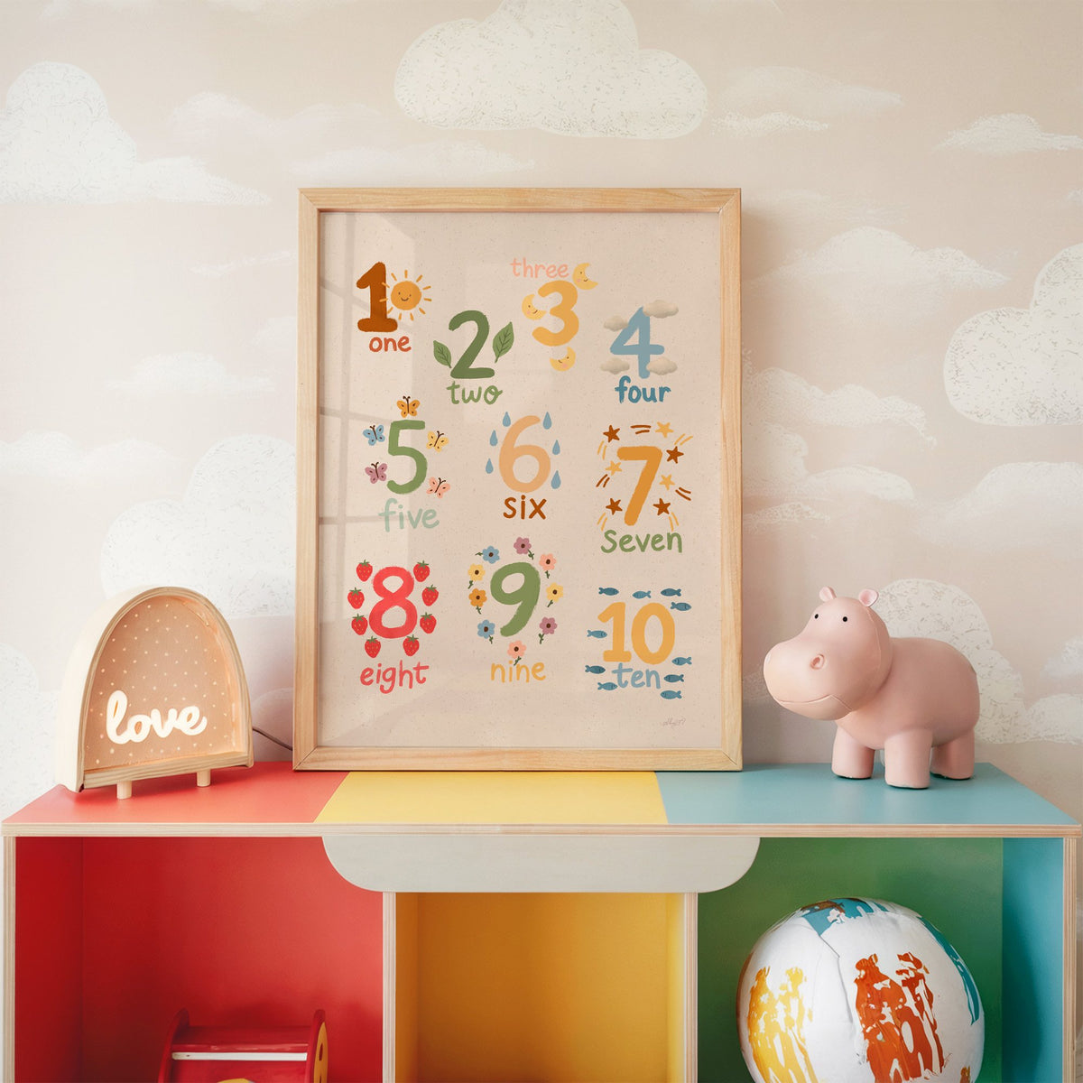 Numbers one to ten counting chart nursery print in an oak frame on a rainbow toy shelf against cloud wallpaper