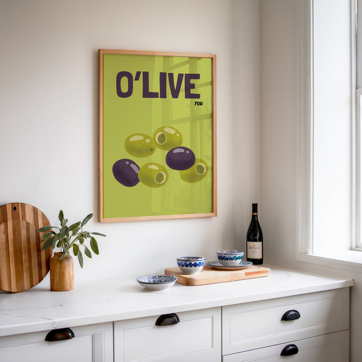 O Live You olives print in an oak frame above a white sideboard with a bottle of wine