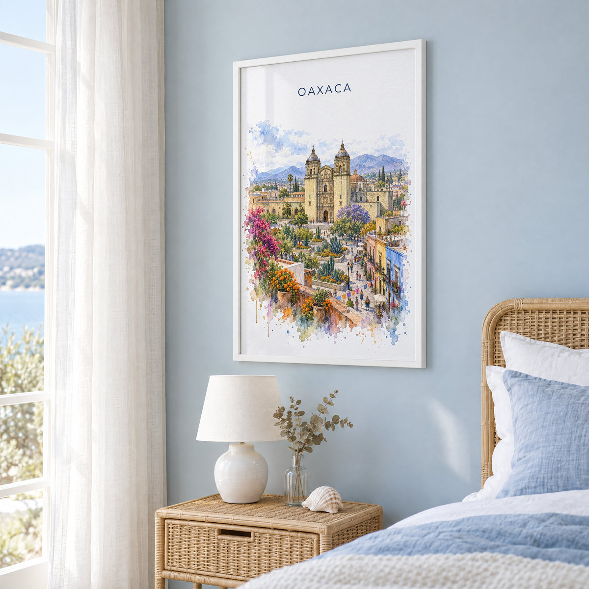 Oaxaca watercolor print in a white frame on a pale blue wall beside a rattan bed in a coastal bedroom