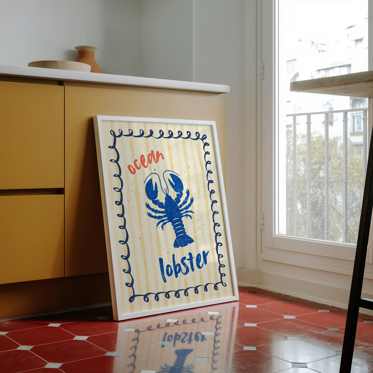 Blue lobster print in a white frame against yellow cabinets on a red tiled floor