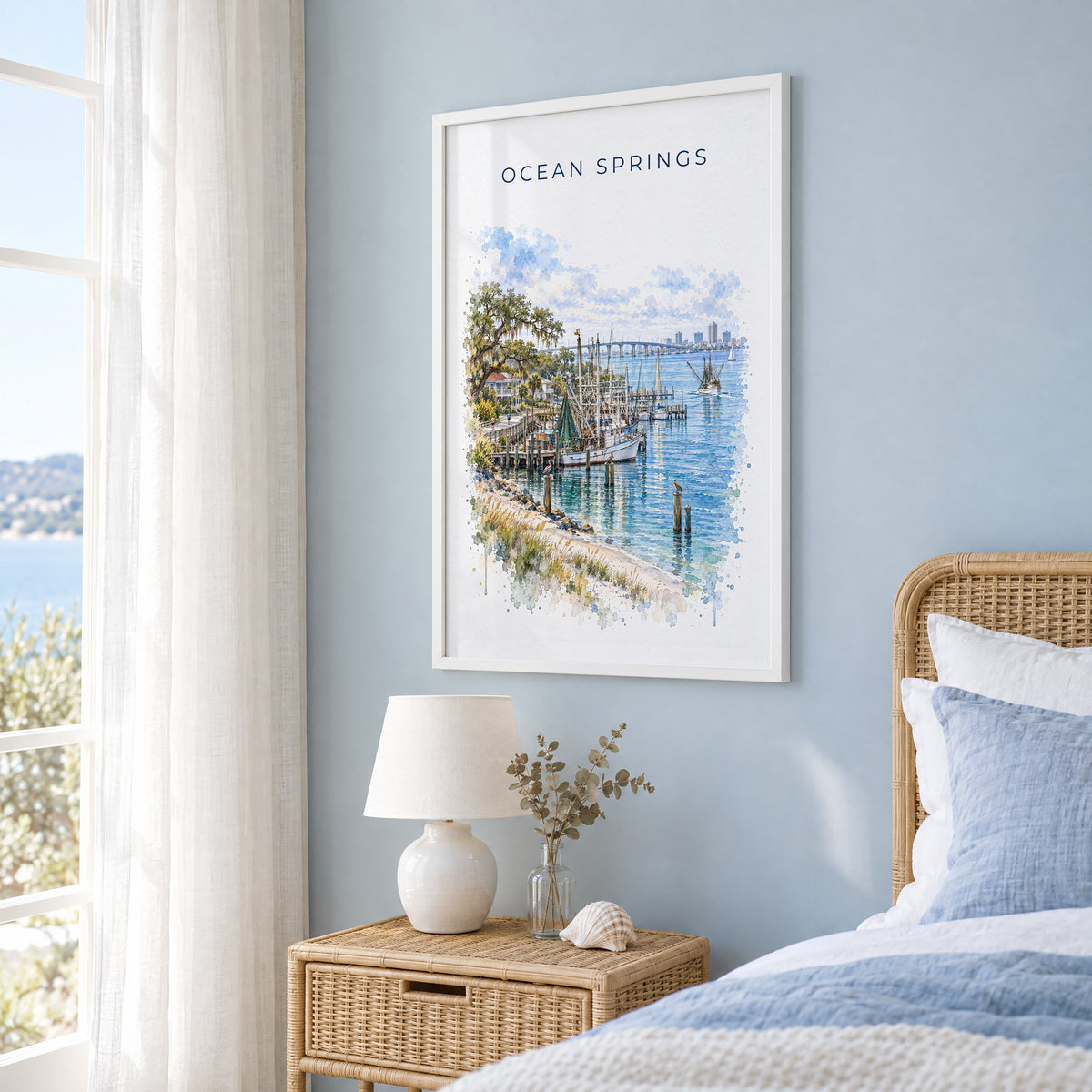 Ocean Springs, Mississippi watercolor print in a white frame on a pale blue wall beside a rattan bed in a coastal bedroom