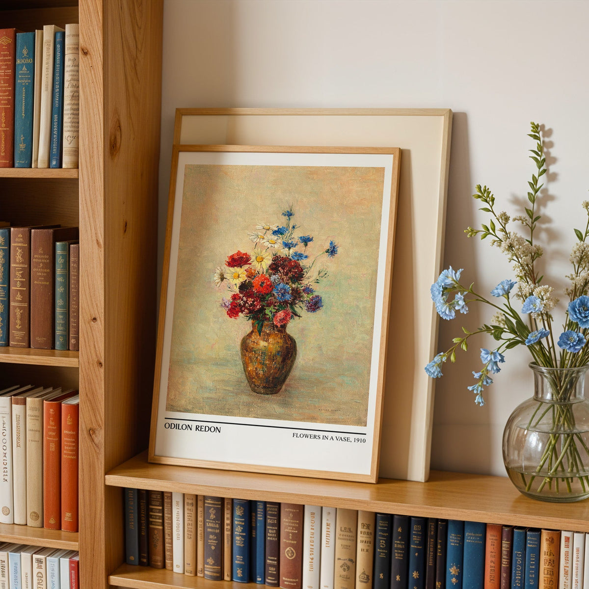 Daisies and cornflowers print in an oak frame leaning on a bookshelf beside flowers