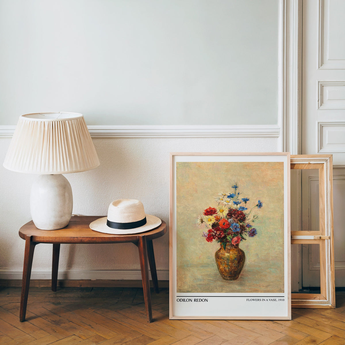 French floral poster in a light wood frame leaning by a bench and hat
