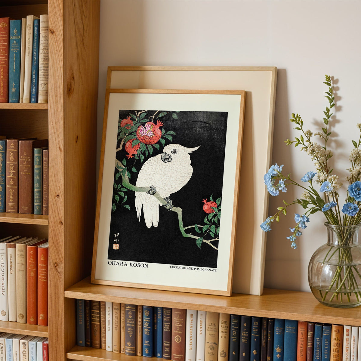 Cockatoo print in an oak frame leaning on a bookshelf beside blue flowers