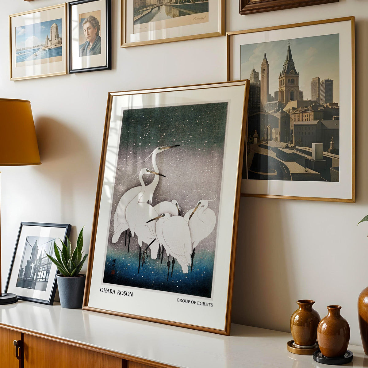 Koson egrets shown in a gold frame leaning on a sideboard in a gallery wall
