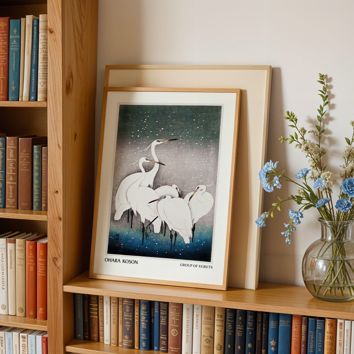 Egret print in an oak frame leaning on a bookshelf beside blue flowers
