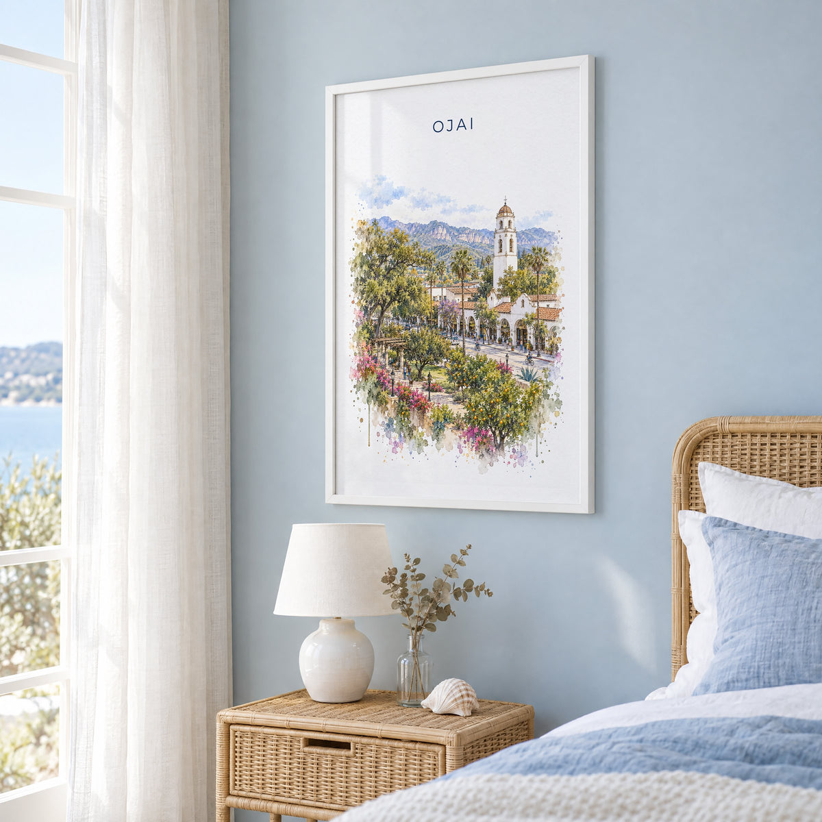 Ojai, California watercolor print in a white frame on a pale blue wall beside a rattan bed in a coastal bedroom