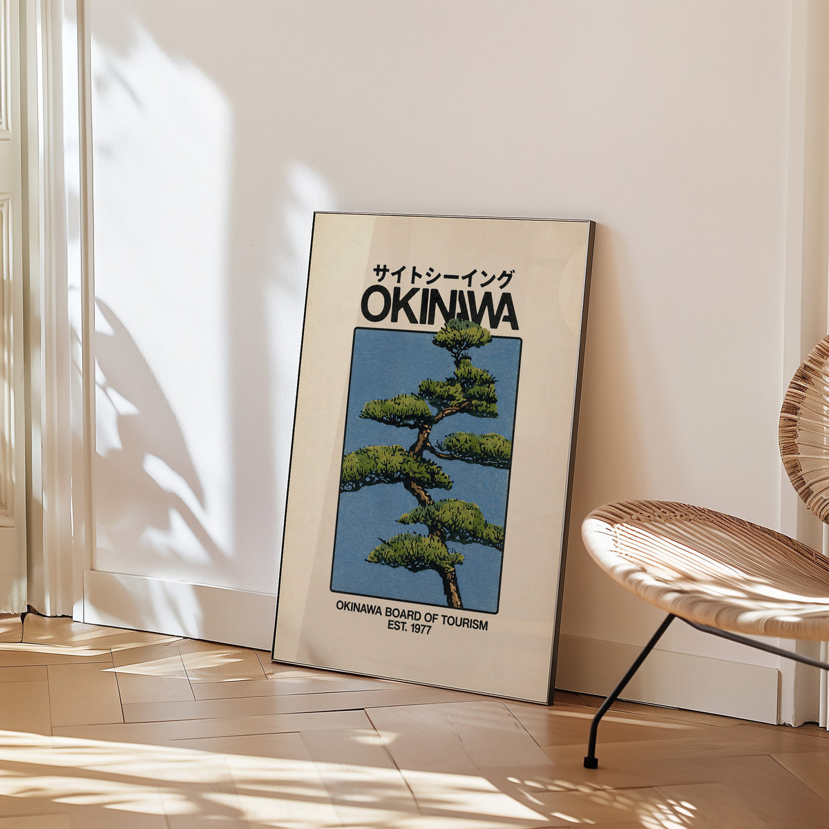 Okinawa pine tree travel poster print in a black frame leaning on a herringbone floor beside a woven rattan chair