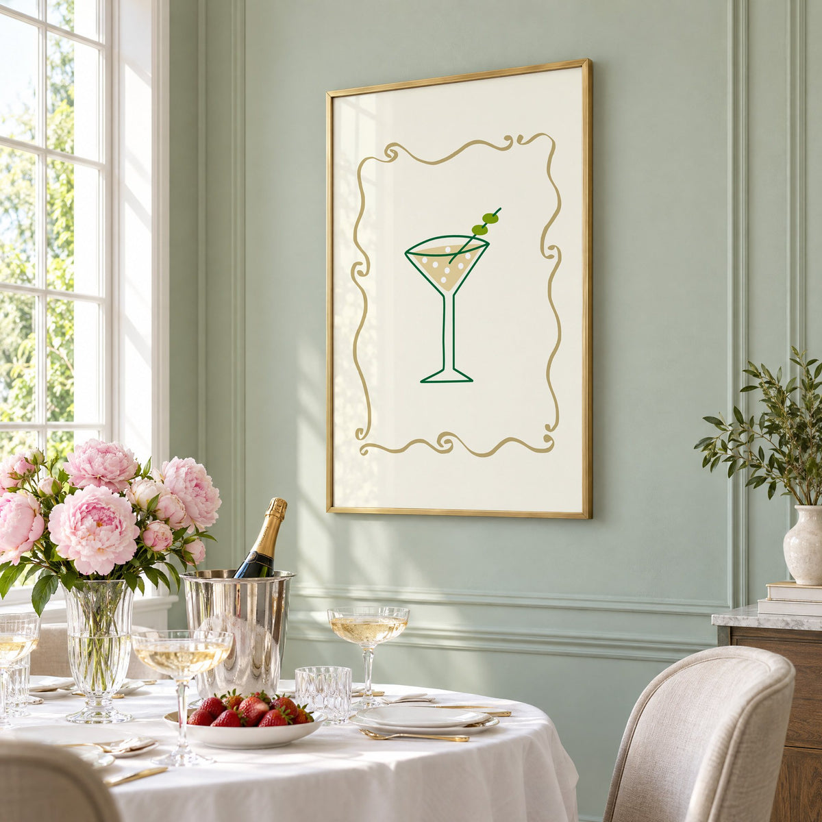 Olive martini in gold scroll frame print in a brass frame on a sage wall above a brunch table with champagne and peonies, seen at an angle